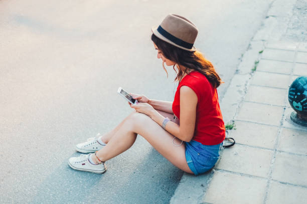 istock Young woman sitting at the sidewalk and texting messaging 1477773446