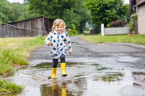 Little toddler girl wearing yellow rain boots, running and walking during sleet on rainy cloudy day. Cute happy child in colorful clothes jumping into puddle, splashing with water, outdoor activity.