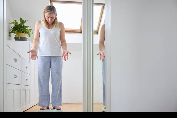 menopausale reife frau, die sich mit gewichtszunahme befasst und im schlafzimmer zu hause auf der waage steht - body concious stock-fotos und bilder