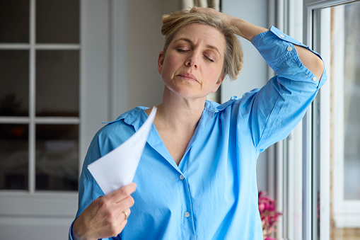 Mujer madura menopáusica que tiene sofocos en casa enfriándose con cartas o documentos photo