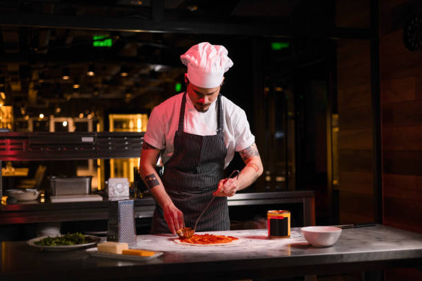 istock Young pizza chef spreading savory sauce on dough in pizzeria. 1477776921