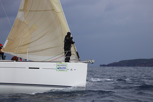 Bodrum,Mugla, Turkey. 05 February 2023: sailor team driving sail boat in motion, sailboat wheeling with water splashes, mountains and seascape on background. Sailboats sail in windy weather in the blue waters of the Aegean Sea, on the shores of the famous holiday destination Bodrum.