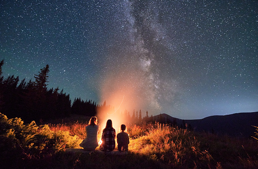 Young family stargazing together in mountains. Three girls warming up near bonfire. Family looking at bright Milky Way in sky.