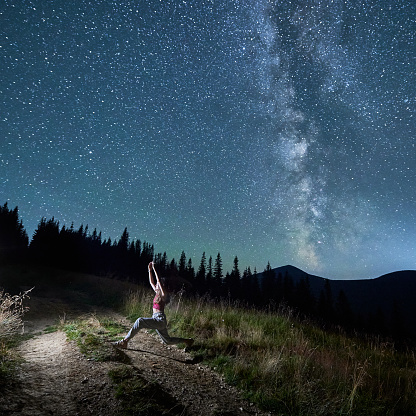 Young girl doing yoga under starry sky. Gymnastic exercises in night mountains. Sporty girl doing yoga pose in moutains on background of Milky Way.