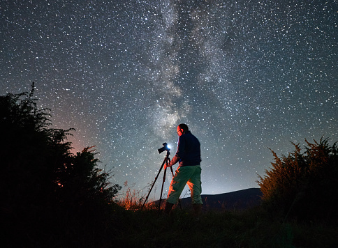 Professional photo shoot of Milky Way in mountains. Male photographer taking photos of starry night in mountains. Back view of man with camera.