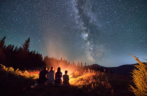 Silhouettes of young family sitting opposite camp fire. Family stargazing in mountains together. Travellers warming up near bonfire.