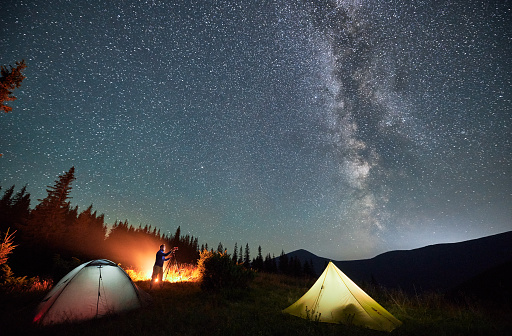 Male photographer taking pictures of Milky way. Man doing photo shoot of starry sky next to his camping. Starry night in mountains.