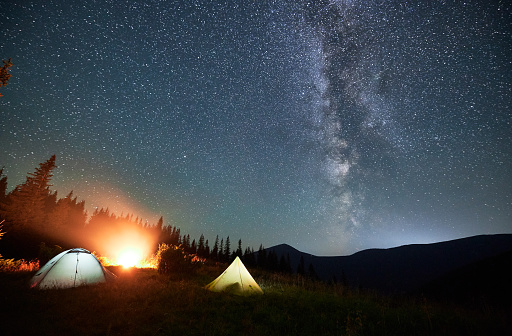 Night camping under sky full of stars and Milky way in the mountains. Starry sky over illuminated tourist tents on hills near forest. Warm light from campfire at dark night.