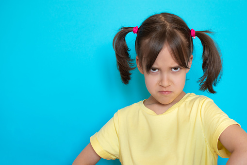 Close-up portrait of upset displeased sulking angry little toddler girl over blue background with copy space.