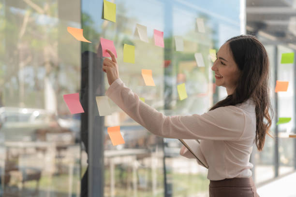 istock Young Asian businesswoman working on a sticky note on board, planning and analyzing business finance and marketing at the office. 1477786469