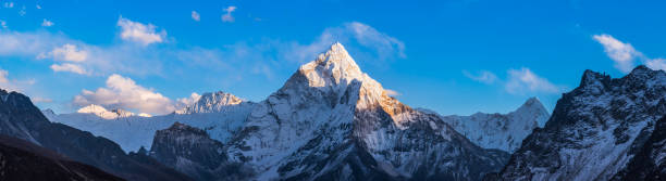 Snowy mountain peak illuminated at sunset wilderness panorama Himalayas Nepal White fluffy clouds in blue high altitude skies over the snow capped peak of Ama Dablam, deep in the Himalayan mountain wilderness of the Everest National Park, Nepal. mountain peak stock pictures, royalty-free photos & images