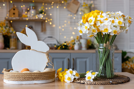 A basket with a wooden white rabbit and natural eggs stands on the table in the kitchen, with Easter decorations nearby glass vase with daffodils flowers. Close up