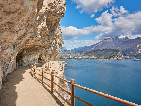 Wide-angle view of a scenic section of Strada del Ponale (Ponale Road), a former highway open to vehicles connecting Lake Ledro and the town of Riva del Garda, nowadays a panoramic route reserved to hikers and cyclists, overlooking the deep blue waters of Lake Garda. The bright, warm light of a spring day, picturesque clouds, high, looming walls of rock, a narrow trail cutting into the side of the mountain. The town of Riva del Garda and the slopes of Monte Baldo can be seen on the background. High level of detail, natural rendition, realistic feel. Developed from RAW.