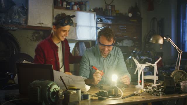 Portrait of Teenage Boy and his Father in Workshop Working Together on Soldering Wires and Circuit Boards for School Science Fair. Man and Son Bonding, Sharing their Hobby, Celebrating their Success