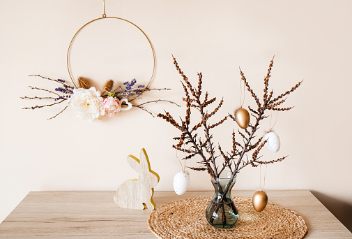 Home Easter decor. Handmade spring round flower garland hanging on wall, tree branches on vase with eggs hanging from it. Nice warm beige earthly tones. Minimal home decor.