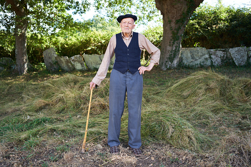 Full length view of an elderly grandfather posing for the camera in his casual attire of trouser, vest, beret, and sneakers, leaning on his walking stick in the Galician countryside.