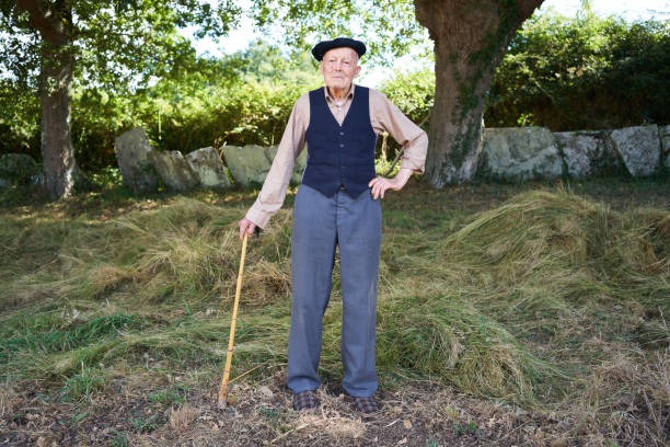 Full length view of an elderly grandfather posing for the camera in his casual attire Full length view of an elderly grandfather posing for the camera in his casual attire of trouser, vest, beret, and sneakers, leaning on his walking stick in the Galician countryside. alternative pose stock pictures, royalty-free photos & images
