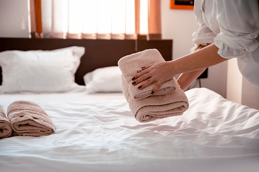 Hotel maid putting set of towel with white flower on the bed. Staff in blue uniform preparing room for hotel guest.