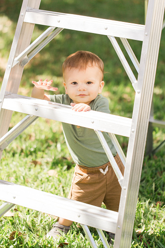 A Cute 15-Month-Old Cuban-American Baby Boy Playing in the Grass & Looking Through a Metal Ladder During Family Yard Work Outdoors on the Front Lawn on a Warm South Florida Spring Day in 2023, Tree Trimming Before Hurricane Season Starts
