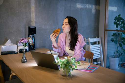 Young woman eating croissant for breakfast and working from home