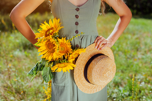 Close up of summer female accessory and clothes. Woman holding straw hat with bouquet of sunflowers outdoors. Rustic boho style