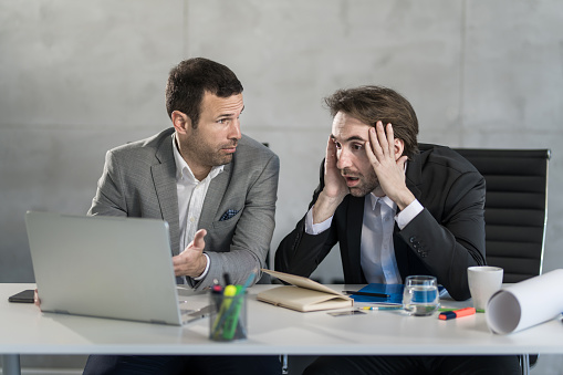 Two shocked businessmen working and seeing bad results on laptop in the office.