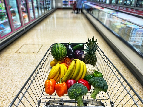 A variety of fresh fruit and vegetables, including bananas, oranges, pineapple and broccoli, peppers and cauliflower in a shopping trolley. Focus on the contents of the cart with the supermarket aisle and shelves defocused beyond.