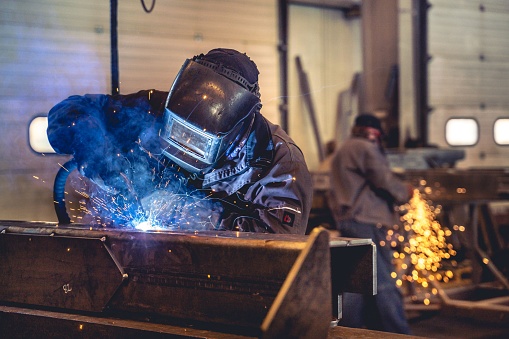 A skilled welder is seen at work in a large industrial factory, using a welding machine to repair a metal plate
