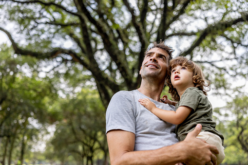 Loving Brazilian father carrying his son at the park while they look up at the trees - lifestyle concepts