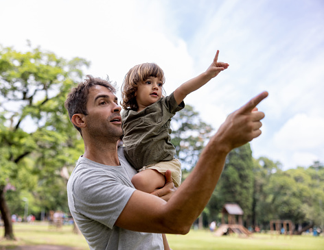 Brazilian father carrying his son while having fun at the park and pointing away - lifestyle concepts