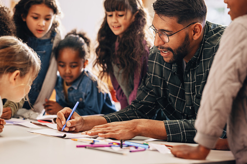 Man teaches his students how to draw in a primary school class. This elementary school educator shows his kids how to use a colouring pencil. Teaching an elementary class in a multiethnic school.