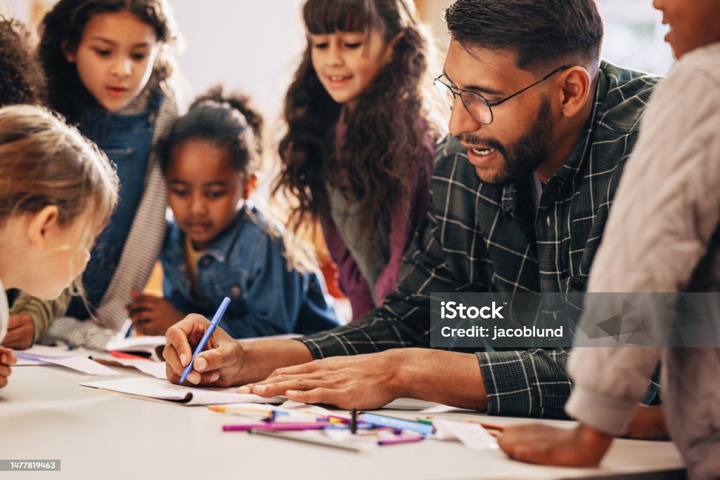 Man teaches his students how to draw in a primary school class Man teaches his students how to draw in a primary school class. This elementary school educator shows his kids how to use a colouring pencil. Teaching an elementary class in a multiethnic school. Teacher Stock Photo