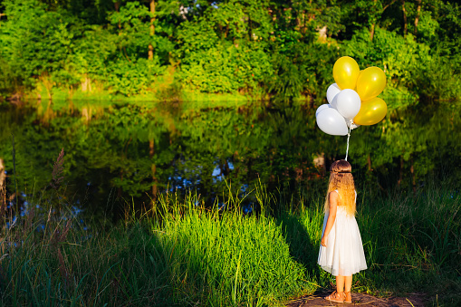 A cute little girl in a white dress with long hair stands with her back, looks at the lake, holding many balloons in her hands.