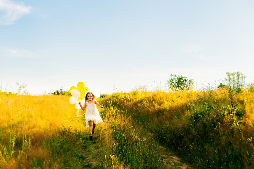 beautiful little girl in a white dress runs on green grass and holds balloons.