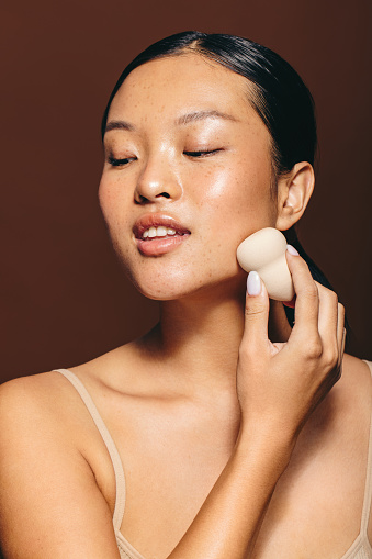 Female with glowing skin applies a facial treatment using a beauty sponge, she gently massages her skin with the blender. With a self-assured expression on her face, she is confident in her skincare routine.