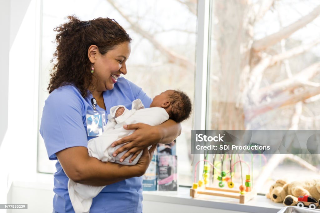 Female nurse looks at baby with smile on her face The mature adult female nurse looks at the baby in her arms with a big smile on her face. Baby - Human Age Stock Photo
