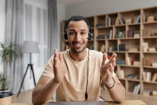 Positive arab freelancer guy in headphones having online video call, speaking to business partner and gesturing at camera, participating in web conference or remote meeting