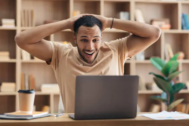 Photo of Omg yes, I did it. Overjoyed arab man using laptop and touching head in excitement, celebrating great news