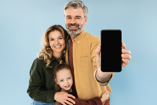 Parents And Daughter Showing Mobile Phone Empty Screen Recommending New App Smiling To Camera Standing On Blue Studio Background. Great Application For Cellphone. Mockup