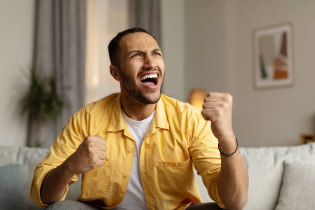 istock Emotional young black man shouting, gesturing YES in joy, celebrating victory, sitting on sofa at home 1477839561