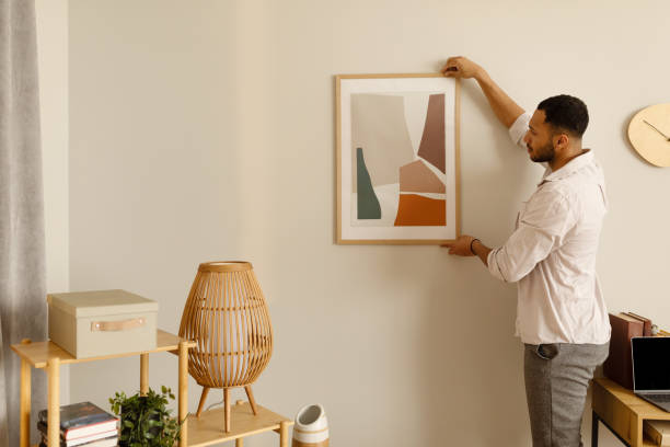 Modern home decor and interior design. Young black man hanging painting, putting picture frame on wall indoors Modern home decor and interior design. Young black man hanging painting, putting picture frame on wall indoors, copy space. Millennial African American guy decorating his apartment hanging art on wall stock pictures, royalty-free photos & images