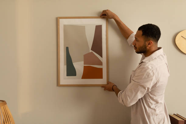 Young black man hanging painting, putting picture frame on wall indoors. Modern home decor and interior design Young black man hanging painting, putting picture frame on wall indoors, copy space. Millennial African American guy decorating his apartment. Modern home decor and interior design hanging art on wall stock pictures, royalty-free photos & images