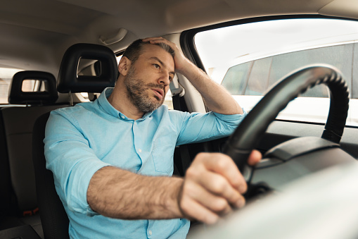 Portrait of upset man driver sitting at front seat and touching his head, riding car. Bired adult guy getting late for work or flight, got stuck in traffic jam, holding streering wheel
