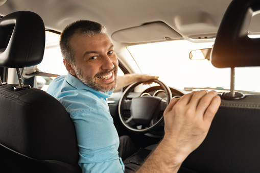 Cheerful man driving new automobile after purchase, sitting on driver's seat of car at dealership store shop. Happy guy looking back at passenger seat and smiling, buying modern auto at showroom
