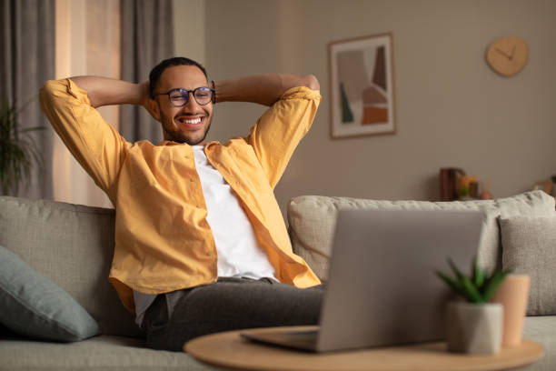istock Smiling young black man with laptop relaxing after working online, sitting on couch with laptop at home, copy space 1477841315