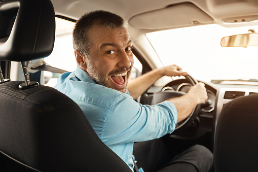 Happy Driver. Portrait of cheerful positive man looking back posing at camera sitting in a car, view from rear passenger seat. Excited emotional guy riding in the city, holding hands on steering wheel