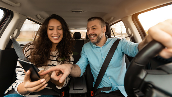 Summer Vacation Concept. Portrait of smiling couple sitting inside luxury car, woman holding cell, cheerful man pointing at mobile phone, showing location, using digital gps map application