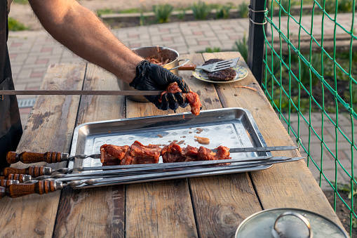 A gloved human hand strings pieces of meat onto a skewer and places them on a metal tray. Outdoor video.