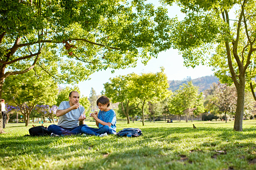 Full length view of mature man sitting cross-legged on grass at public park with 11 year old son, enjoying weekend leisure together.