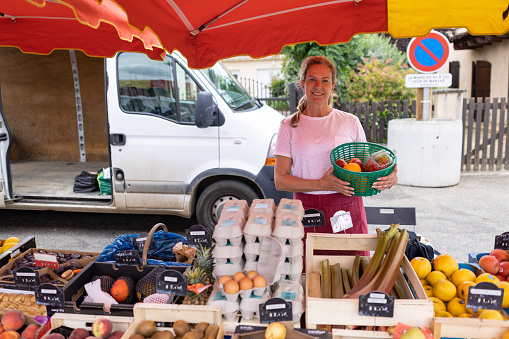 A portrait front view of a female greengrocer standing proudly at her market stall in a small village called Mosaic in Toulouse in the south of France. She is holding a basket of locally grown and produced organic fruit and veg.